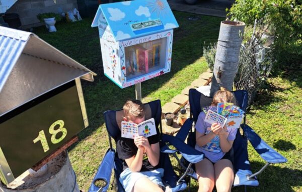 Children reading books next to a Street Library. Their faces are obscured by their books.